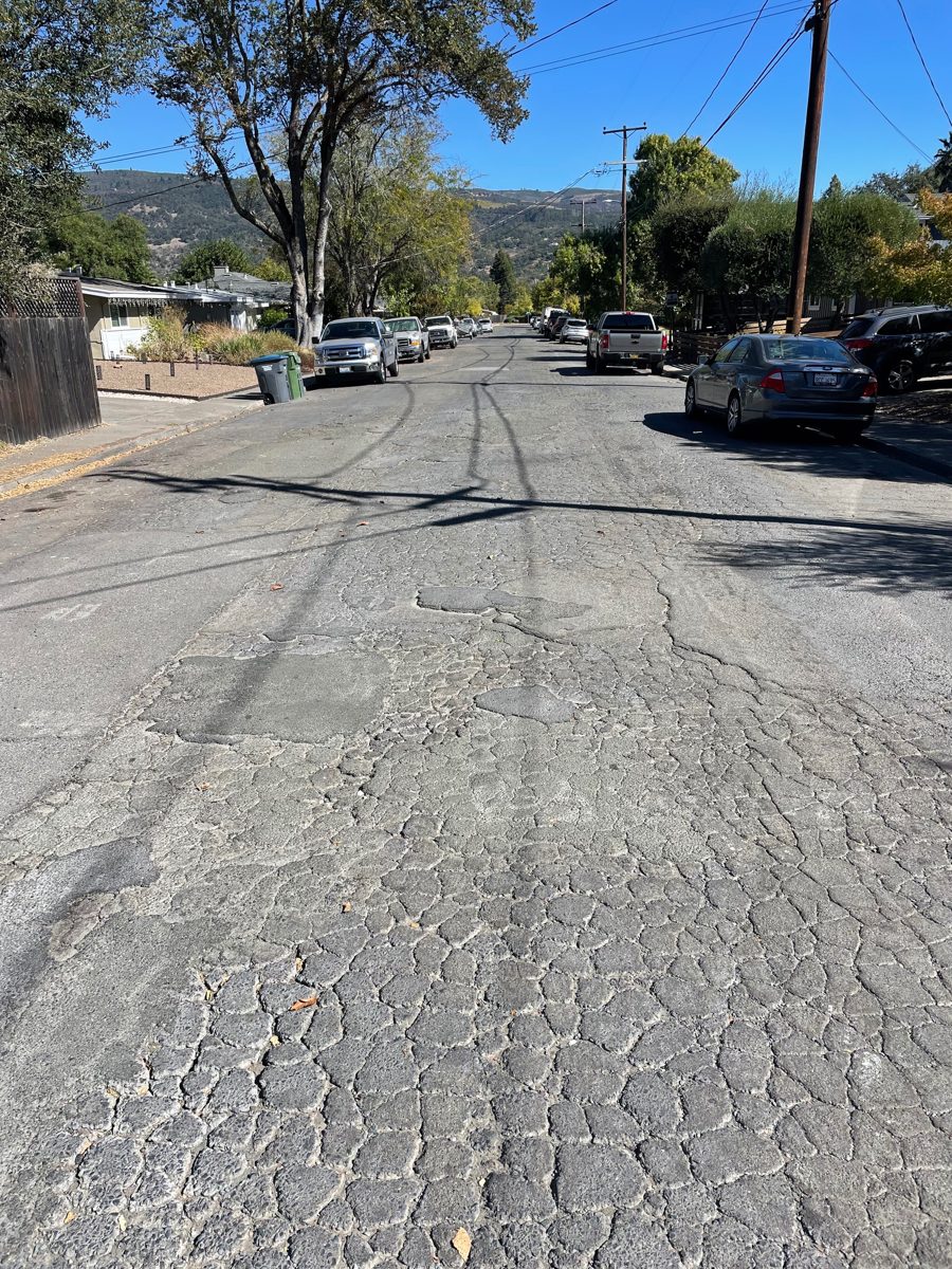 A long residential street with cracking in a tight block pattern across both lanes and patched sections