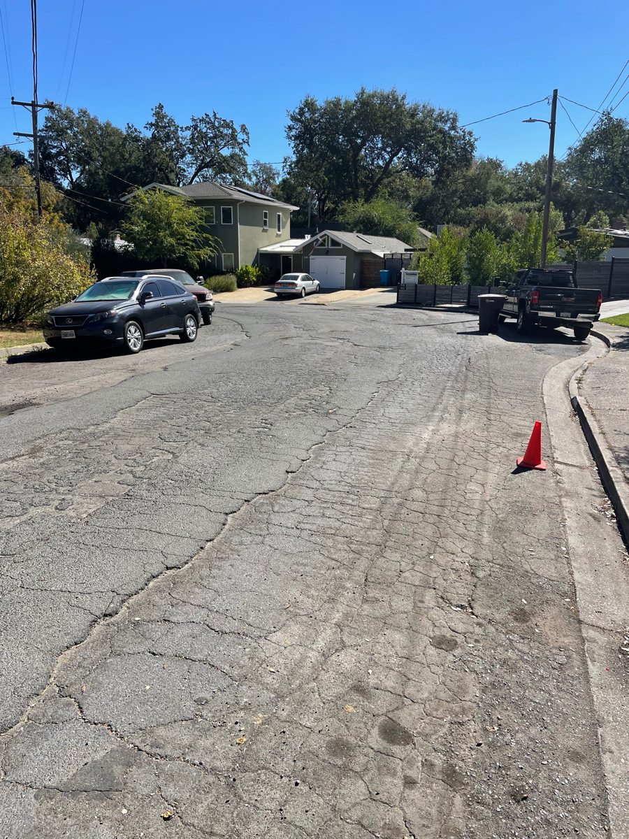 Curving street with cracks running its length and an orange traffic cone at the curb
